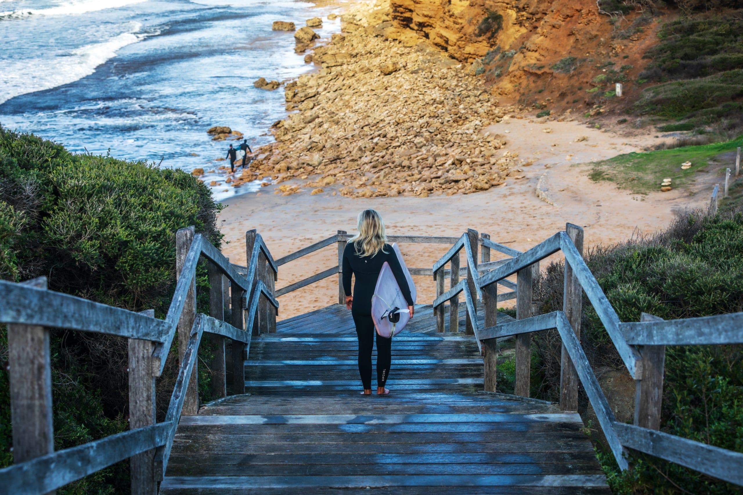 A surfer carrying a longboard down wooden beach stairs toward the water