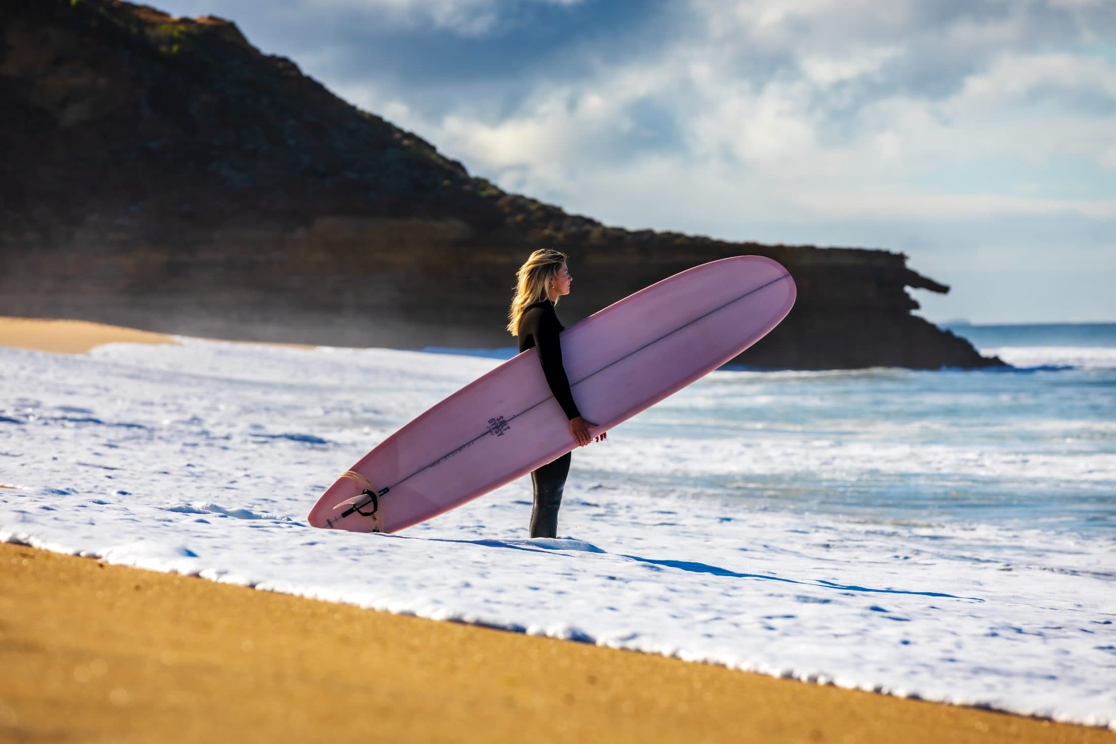 A surfer with a longboard standing in the shorebreak foam below a dark headland